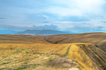 Golden hills and fields with a winding path lead toward a distant mountain range under a vast blue sky with clouds. This peaceful landscape highlights the beauty of nature's wide-open spaces.

