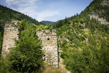Fototapeta premium Summer view of the ruins of Gogia Fortress against a mountainous backdrop in Borjomi, Georgia. The ancient stone walls are surrounded by green trees, creating a striking contrast with the landscape