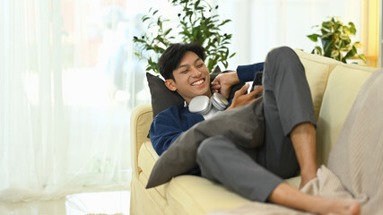 Obraz premium Cheerful young man using smartphone and relaxing on sofa