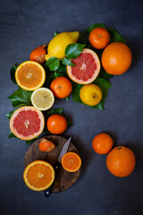 Aerial view of sliced ​​oranges, tangerines, grapefruits and lemons with green leaves and knife in circle on grey background, vertical with copy space