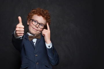 Clever school kid young boy in glasses showing thumb up on black background with copy space