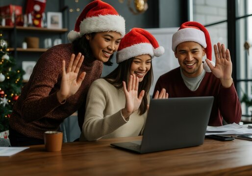 Asian coworkers wearing santa hats waving at laptop during a video call in the office during christmas holidays