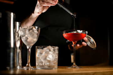 Bartender hand holds a berry with tweezers above tall stemmed wine glass filled with brown cocktail