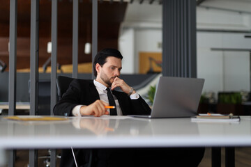 A man in a suit is sitting at a desk with a laptop and a pen. He is looking at the laptop screen...