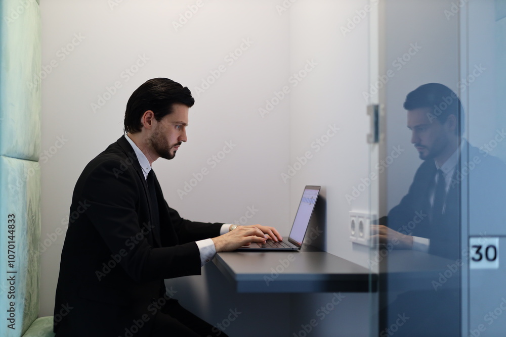 Poster a man in a suit is typing on a laptop in a cubicle. the cubicle is a small, enclosed space with a gl