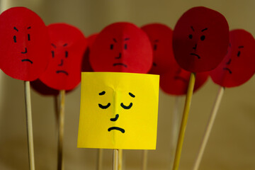 Close up of yellow paper with sad face surrounded by angry faces, concept of social rejection and...