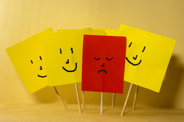 Red paper with sad depressed face surrounded by yellow smiling papers, concept of depression and...