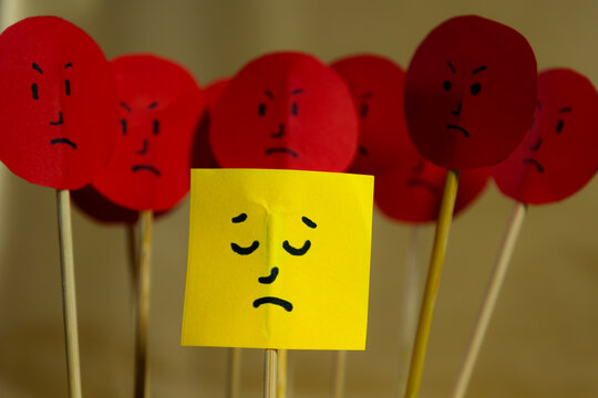 Close up of yellow paper with sad face surrounded by angry faces, concept of social rejection and discrimination