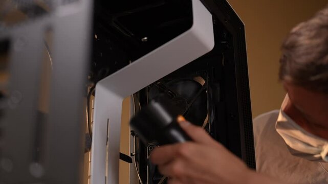 Technician in protective face mask using air blower to clean computer case, eliminating dust for improved performance. Preventing computer overheating, male using mini vacuum to clean case slow motion