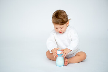 A baby boy with a bottle of milk on a white isolated background with a place for text is sitting in white knitted clothes. A small happy child drinks milk, space for text