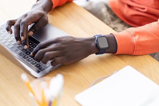 A close-up view focusing on the hands of a man wearing a smartwatch as he types on a laptop, with an orange sleeve visible, set against a wooden table