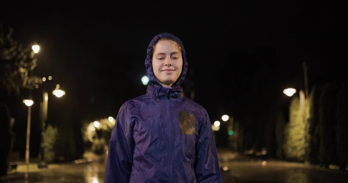Portrait of a smiling runner standing outdoors on a rainy night wearing a waterproof jacket and enjoying the serene ambience of the street illuminated by soft lights.