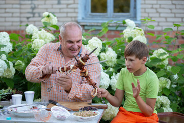 A grandfather and his grandson are happily eating shashlik from a skewer and chatting in the yard of a village house. A man and a boy are eating together.