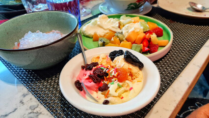High Angle View of Delicious Melting Indonesian Coconut Milk Ice Cream Es Puter In Strawberry, Vanilla Flavors Topped With Dates Orange Raisins Colorful Sprinkles Served In White Bowl With Spoon In It