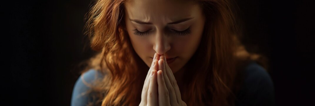 A woman is praying with her hands clasped together. She has red hair and is looking down