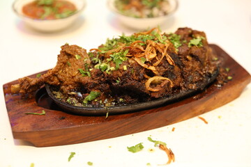 Sizzling mutton curry is served on a wooden board, topped with crispy onions and fresh herbs, with chutney and salad in the background.