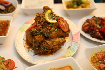 Golden-brown chicken garnished with lemon slices and fresh herbs, alongside various vibrant side dishes presented in white bowls, like rice, a spicy curry, and tangy vegetables.