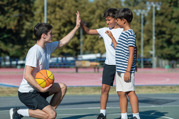 Obraz premium Coach and two young boys exchange a high five during outdoor basketball practice, capturing a moment of teamwork, encouragement, and active participation in youth sports