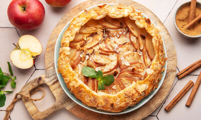 Homemade apple tart with almond toppings on a wooden board surrounded by fresh apples and cinnamon