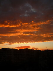 Bright unusual sunset city landscape orange pink rays above cityscape. Brutalist panel buildings appear as black silhouettes against a fiery orange-yellow sky dramatic lighting dark rain storm clouds