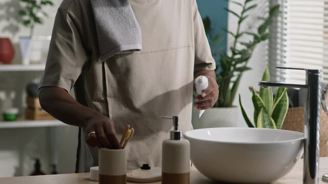 Cropped shot of Black man with towel on his shoulder standing by ceramic white sink in bright sunlit bathroom, squeezing toothpaste on his eco-friendly bamboo toothbrush