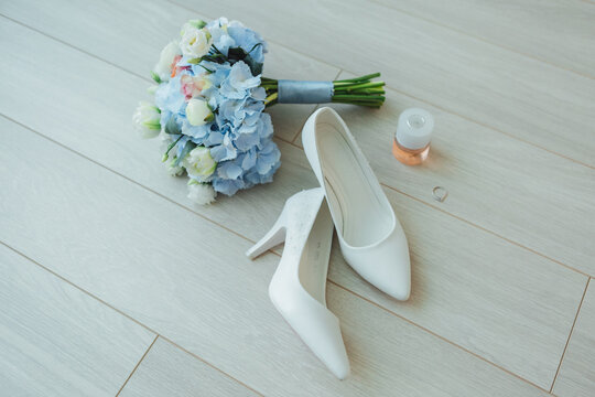 Wedding flat lay with white shoes, blue bouquet, perfume, and a ring on wooden floor.