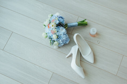 Wedding flat lay with white shoes, blue bouquet, perfume, and a ring on wooden floor.
