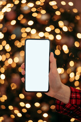 Close-up smartphone with empty screen mockup on holiday background with Christmas tree, customer holding phone in hand