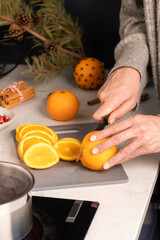 A woman cuts orange slices to make mulled wine near a stove with a saucepan. 