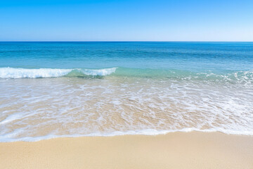 Serene view of gentle ocean waves softly lapping onto a smooth sandy shore with clear turquoise water extending to the horizon beneath a clear blue sky