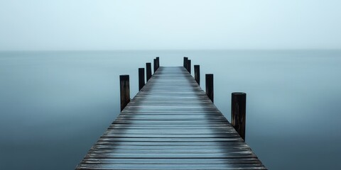 Obraz premium Misty wooden pier extends into calm water under gray sky during early morning hours, creating a serene and tranquil atmosphere
