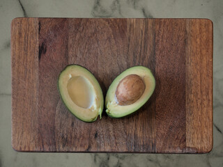 An avocado split in half on a wooden board, on a stone kitchen top