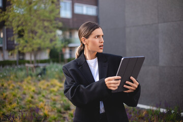 Doubtful woman with ponytail wear suit blazer hold tablet and look mad puzzled, angry. Girl stand near office at city, look annoyed.
