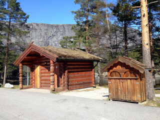 A rest area with toilets and rubbish bins built into traditional-looking cabins by a road in Norway.
