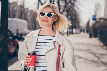 Photo of pretty young blonde woman hold coffee paper cup going wear stylish trench coat walk...