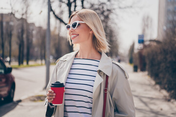 Photo of positive young blonde girl wondered drink coffee wear trendy trench coat walk city sunny spring weather outside