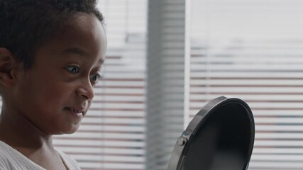Medium closeup of young Black boy gazing into round bathroom mirror, playfully exploring his reflection with curiosity - Powered by Adobe