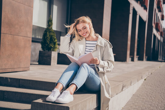 Full body photo of pretty young blonde woman reading book sit concrete border wear stylish trench coat walk outside urban city streets
