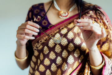Dressed in beautiful saree, Indian woman delicately putting traditional jewellery necklace getting ready for ethnic festive celebration. Indian culture, ritual holiday in modern life. Selective focus.