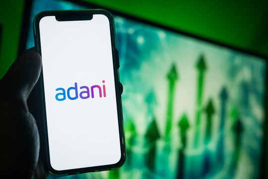 Hand Holding Phone with Adani (ADHI) Logo and Monitor in the Background Showing Upward Green Arrows Suggesting Positive Stock Movement