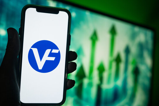 Hand Holding Phone with V.F. Corporation (VFC) Logo and Monitor in the Background Showing Green Arrows Pointing Up Suggesting Stock Growth