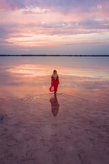 Aerial of a young woman in red dress walking in the water of a unique pink salt lake. Sunset at lake Bursol with beautiful reflections on calm water surface. Stunning scenery