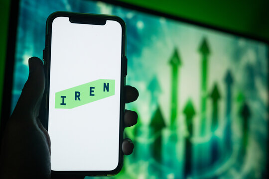 Hand Holding Phone with Iren (IREN) Logo and Monitor in the Background Showing Green Arrows Pointing Up Suggesting Stock Increase