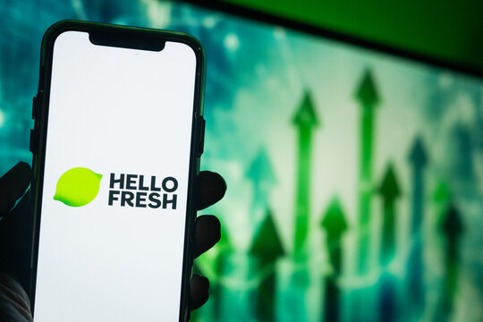 Hand Holding Phone with HelloFresh (HFG) Logo and Monitor in the Background Showing Upward Green Arrows Suggesting Positive Stock Movement