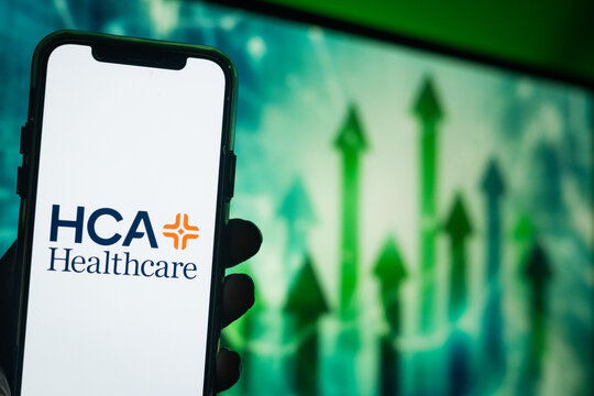 Hand Holding Phone with HCA Healthcare (HCA) Logo and Monitor in the Background Showing Upward Green Arrows Suggesting Positive Stock Movement