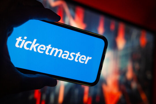 Hand Holding Phone with Ticketmaster (LYV) Logo and Monitor in the Background Showing Red Arrows Pointing Down Suggesting Stock Decline