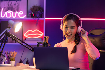 Smiling creative young beautiful Asian influencer talking in channel with positive podcast speech at night time, using laptop for presentation online at pink neon light modern studio. Stratagem.