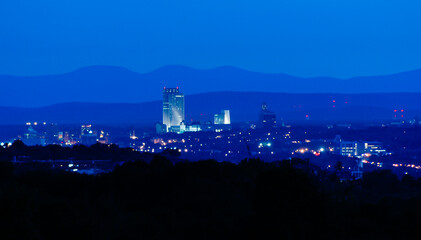 The illuminated skyline of Albany, NY, at dusk, with the Helderberg and Catskill mountain ranges in background. Deep blue evening.
