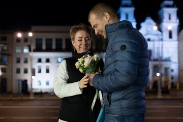 A couple walks through the city at night, the man presenting his wife with a bouquet of flowers