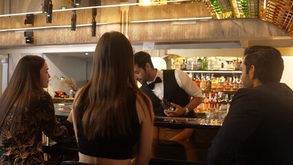 Smart bartender with VIP customer at luxurious bar greeting new beautiful guest at counter in engaging lively conversation at back view in lighting atmosphere at lighting warm nightclub. Vinosity.